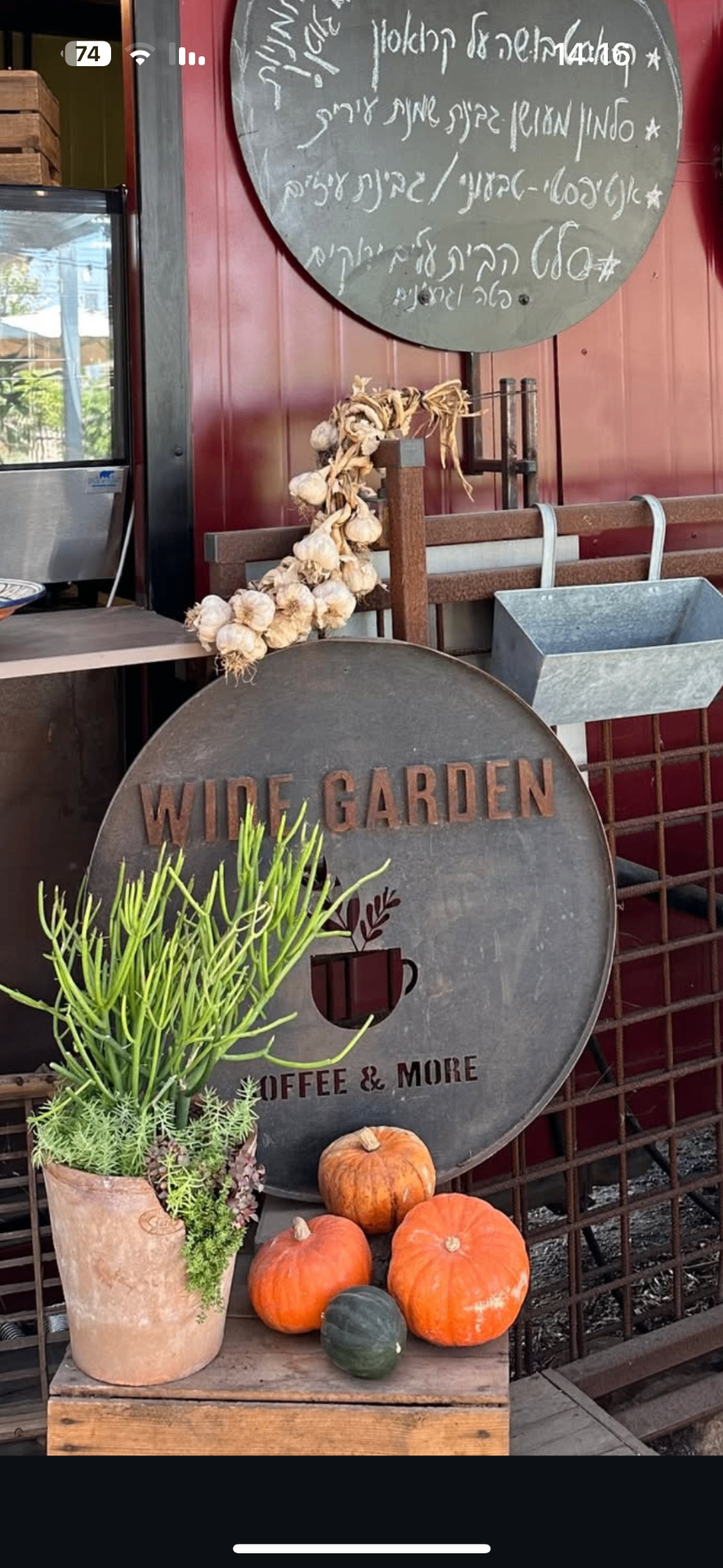 שלט מתכת עגול „WIDE GARDEN Coffee & More” עם אותיות חלודות, ליד עציץ ודלעות