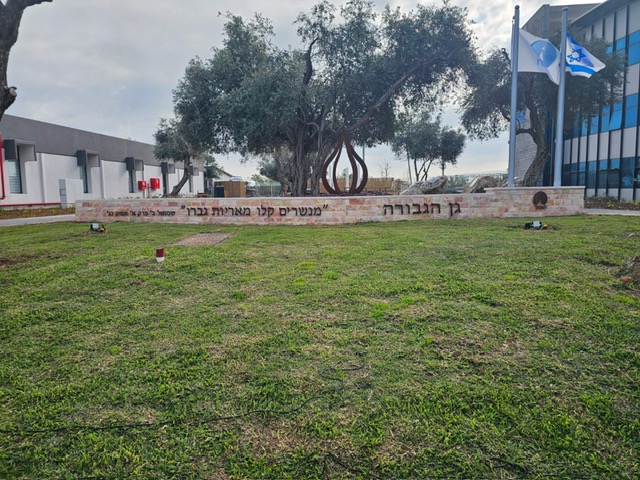 מבט רחב על גן הגבורה עם קיר אבן, כיתוב מתכת, עצי זית ודגלי ישראל
